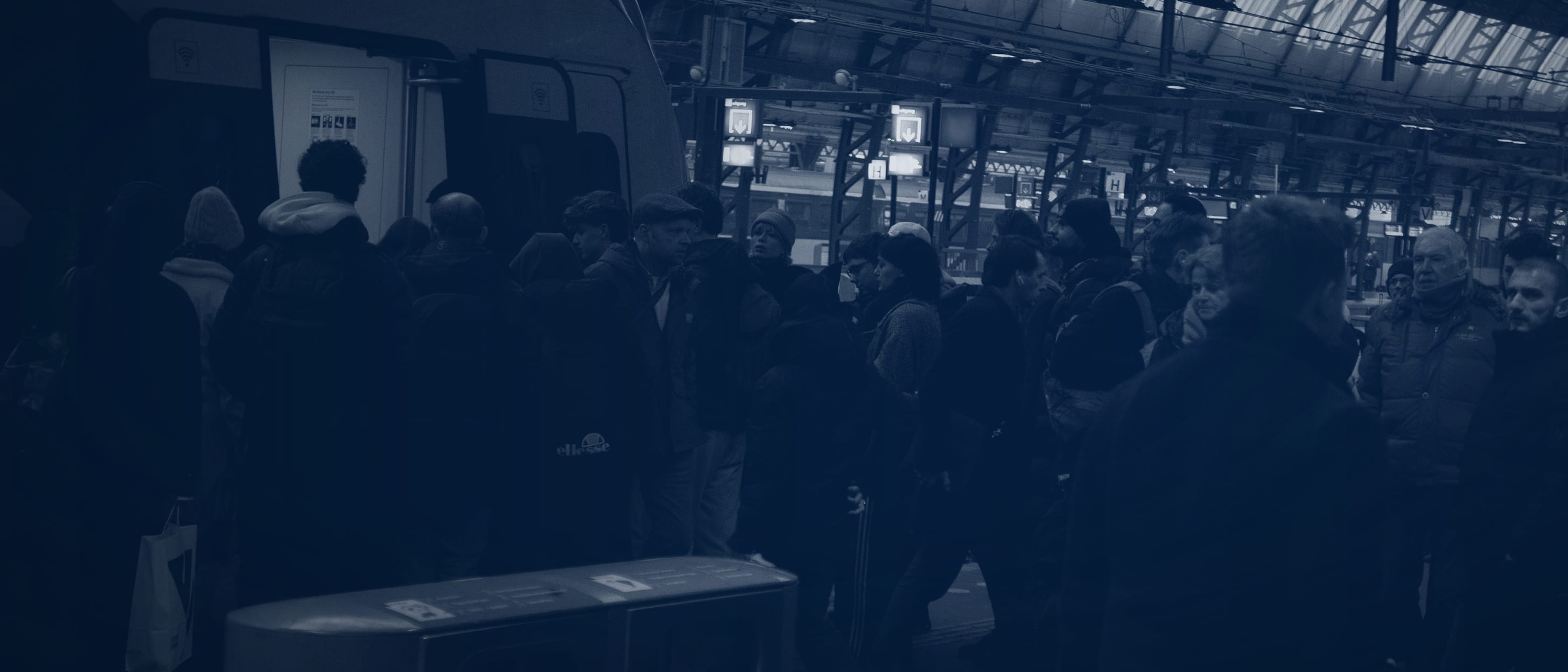 Passengers seen from behind boarding a train on a platform beneath a station roof - no faces are visible.