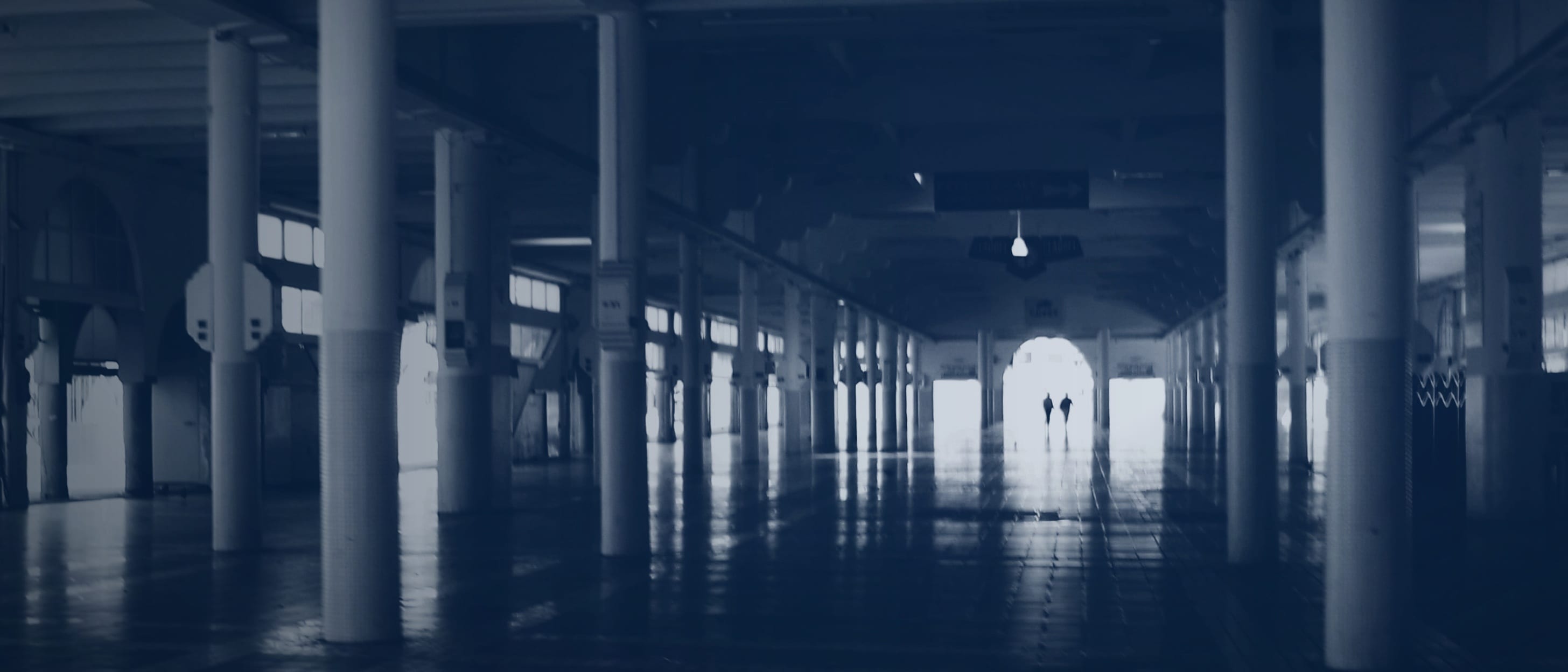 Fast leere Säulenhalle mit glänzendem Boden, am hellen Ende zwei kleine Silhouetten - einzelne Personen sind nicht erkennbar.