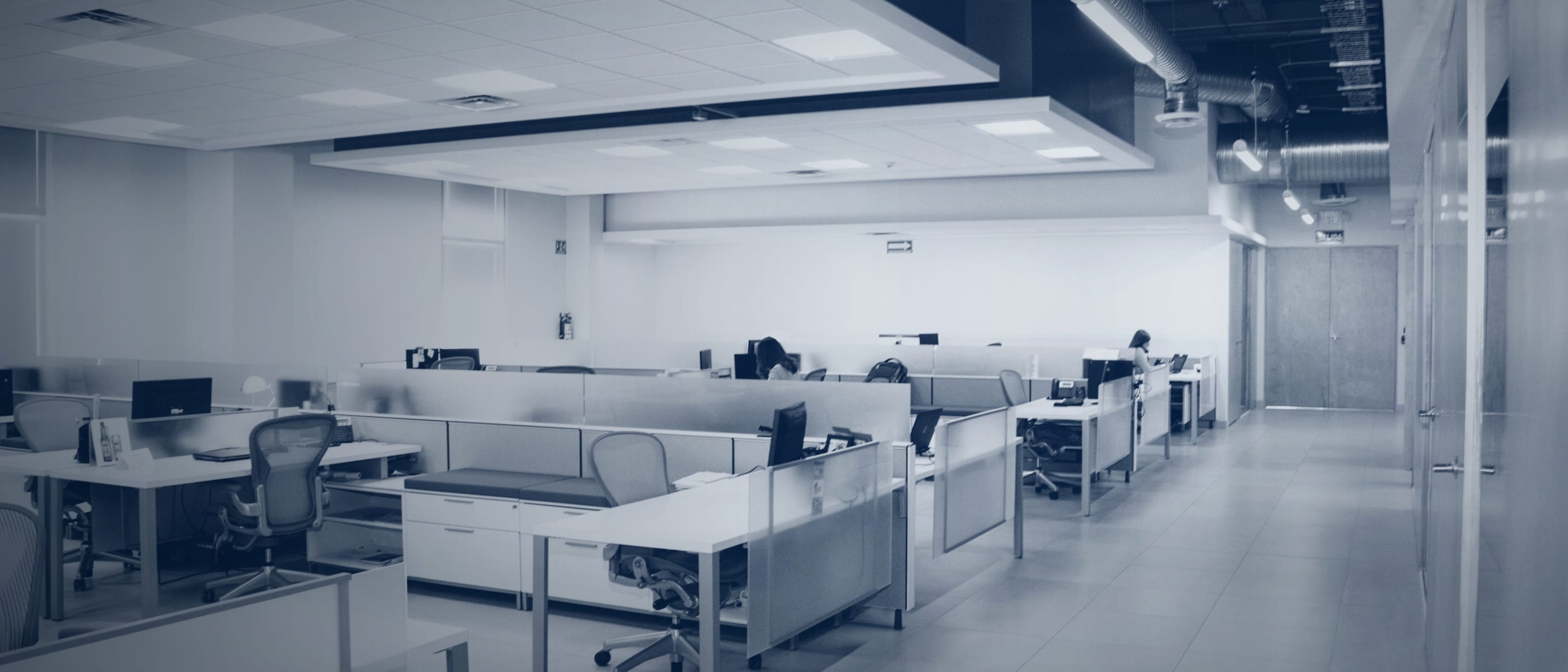 An open-plan office with many empty desks and chairs, two people at their tables far in the background - nobody is identifiable.