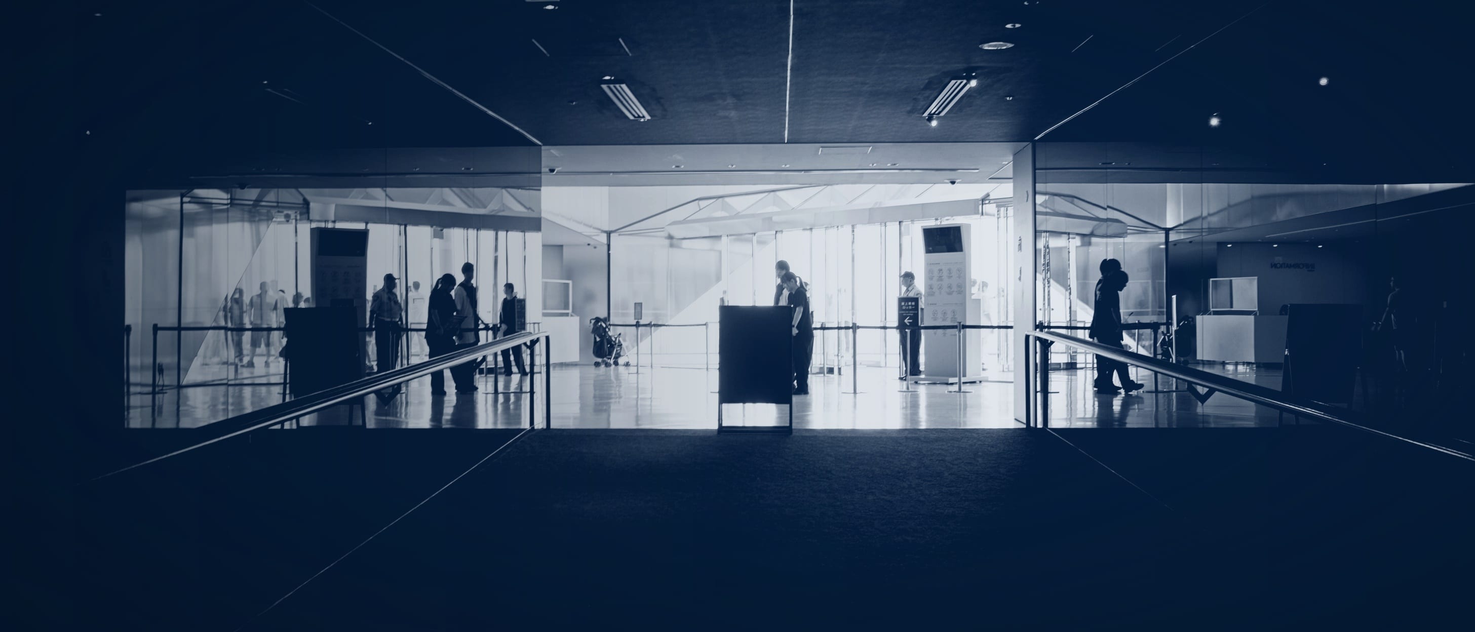 Eingang einer hell erleuchteten Halle am Ende eines dunklen Gangs, Menschen als Silhouetten hinter der Glasfront - einzelne Personen sind nicht erkennbar.