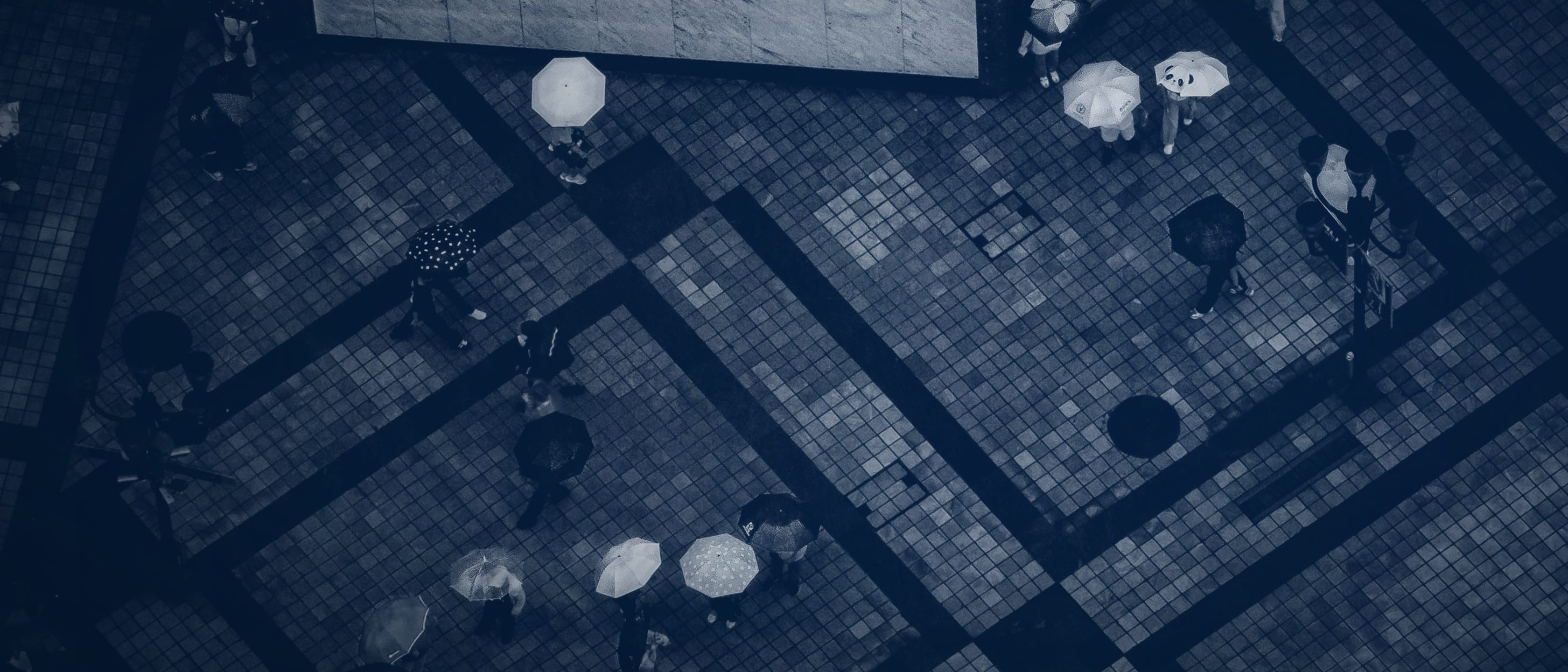 People with umbrellas on a patterned square, seen from high above - no individual is identifiable.