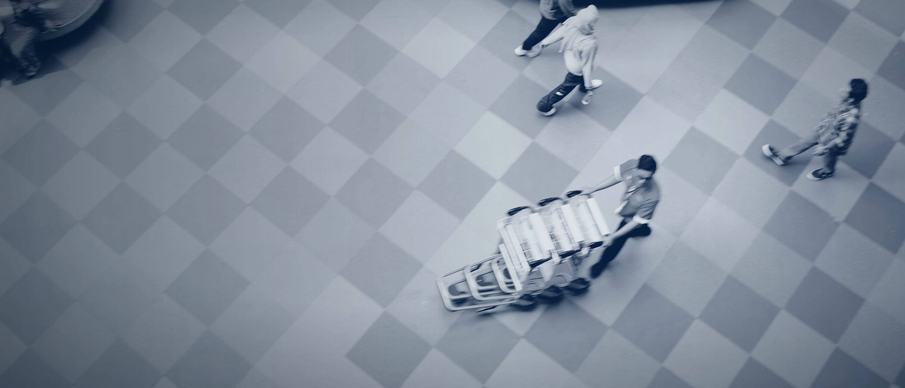 A tiled shopping arcade floor from above: people walking in different directions, one pushing a stack of trolleys - nobody is identifiable.