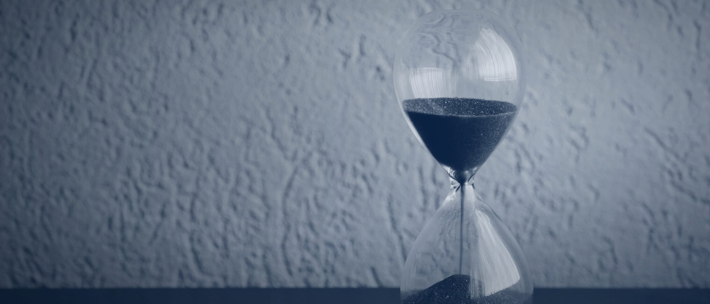 Eine Sanduhr mit dunklem Sand vor einer hellen, rauen Wand auf einem dunklen Tisch - niemand ist abgebildet.