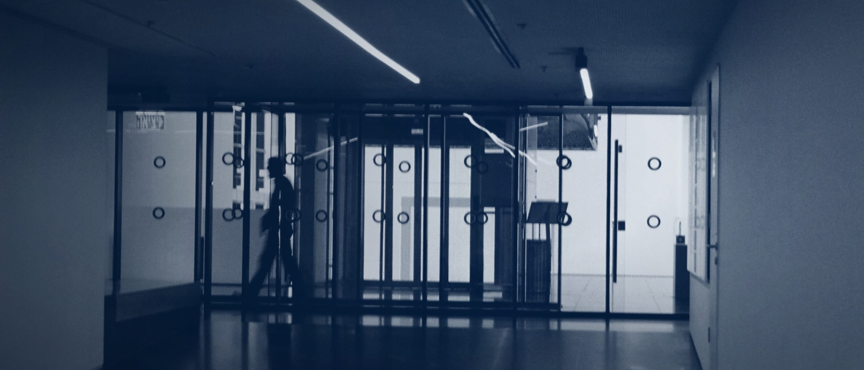 Eine Silhouette geht hinter einer langen Reihe von Glastüren vorbei, der Raum davor liegt im Halbdunkel - niemand ist erkennbar.